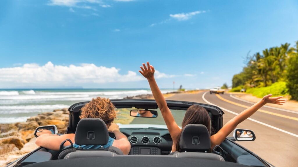 Road Trip Auto Urlaub glückliches Paar fahren Cabrio Auto auf Sommerreise Hawaii Urlaub. Frau mit erhobenen Armen, die sich amüsiert, junger Mann am Steuer